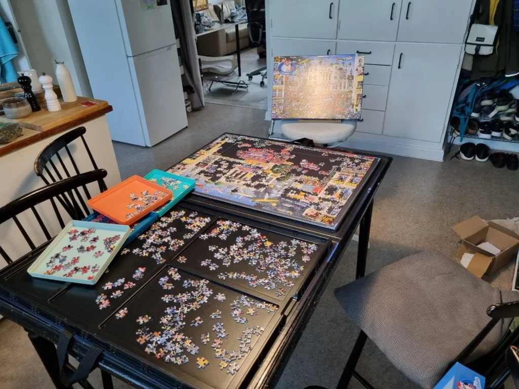Real home table setup for working on an inexpensive jigsaw puzzle, with sorting trays, scattered pieces, and a large completed section visible.