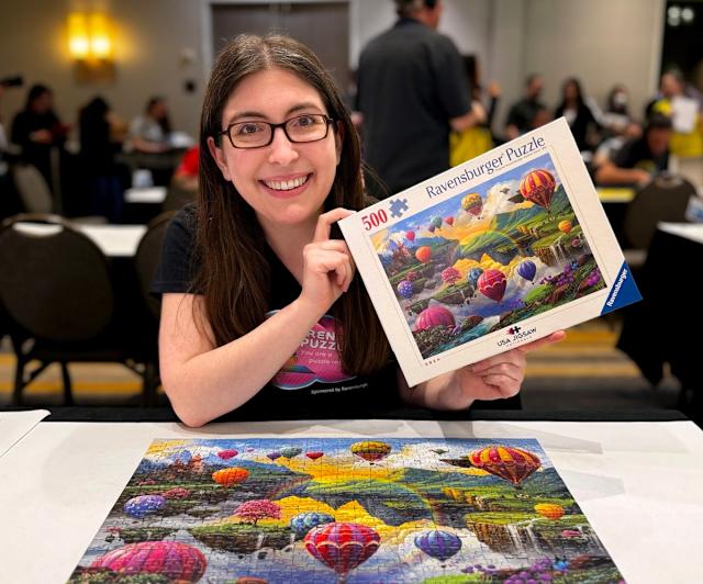 Woman smiling while holding a colorful budget jigsaw puzzle box and showing a completed vibrant puzzle scene, representing satisfying cheap puzzles in 2026.